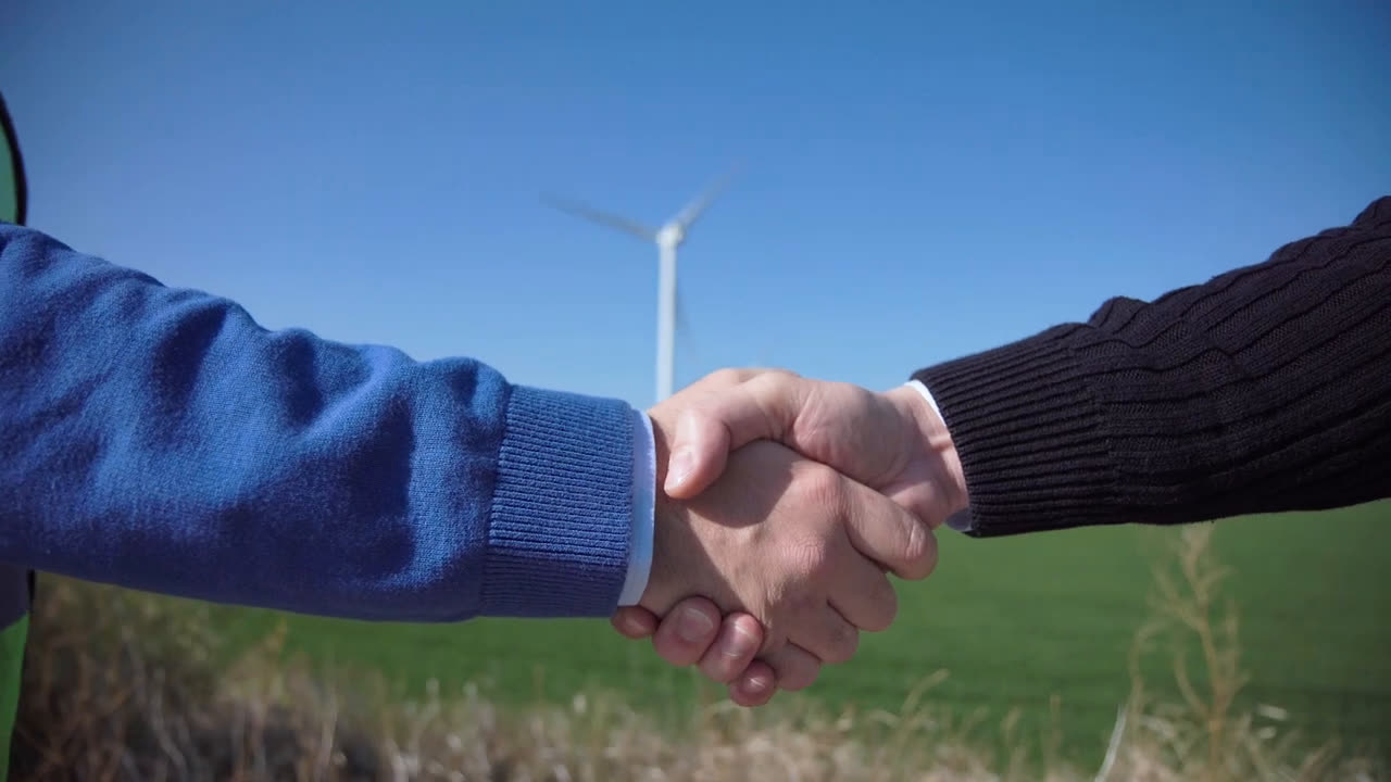 Fotograma de la pieza de Salud Planetaria: dos personas se dan la mano en un campo con un aerogenerador al fondo